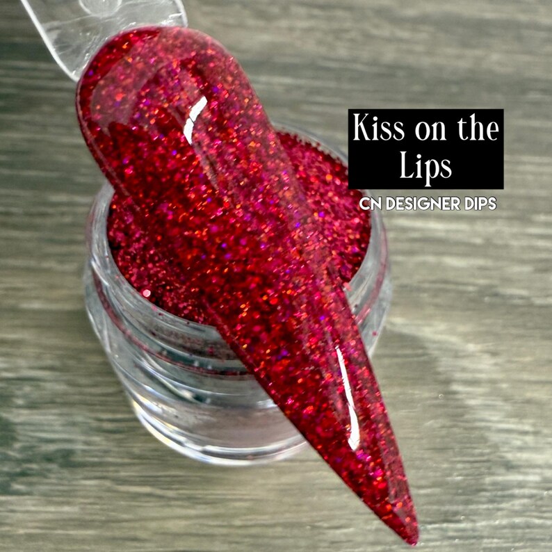 Puede incluir: Primer plano de una u&ntilde;a sumergida en purpurina roja vibrante. La u&ntilde;a se sostiene sobre un peque&ntilde;o tarro transparente lleno de la misma purpurina. El texto "Kiss on the Lips" y "CN DESIGNER DIPS" son visibles.