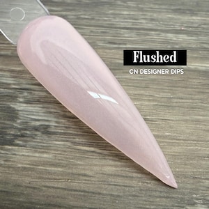 May include: A pale pink nail dip sample with a glossy finish. The sample is labeled "Flushed" by CN Designer Dips. The nail is long and pointed, with a smooth, even surface. The background is a wooden surface.