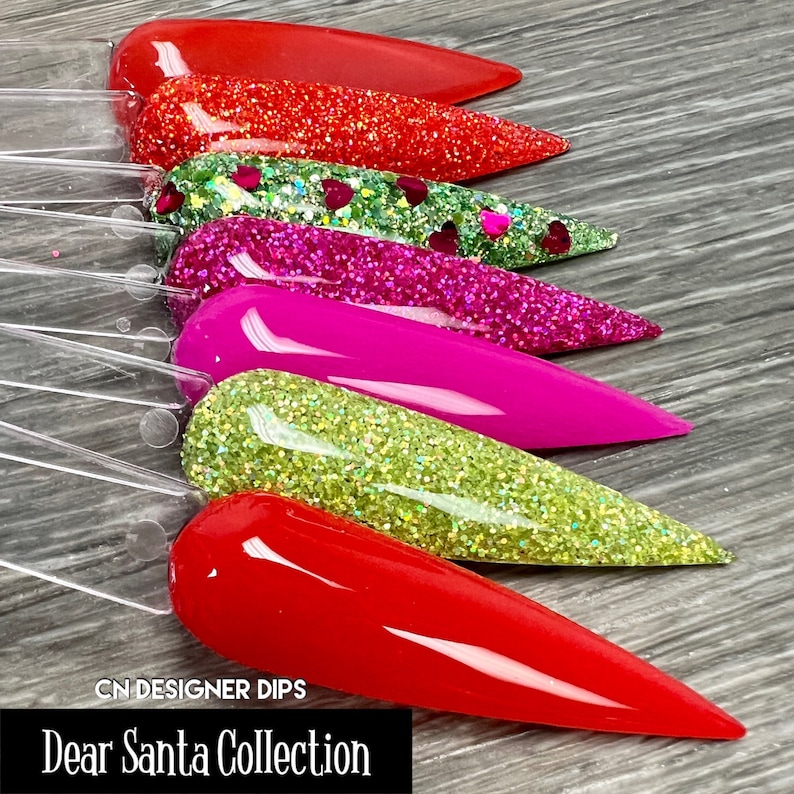 May include: Six different colors of glitter nail dip powder in a long, pointed shape. The colors are red, green with red hearts, pink, green, red, and red. The text "CN DESIGNER DIPS Dear Santa Collection" is at the bottom of the image.