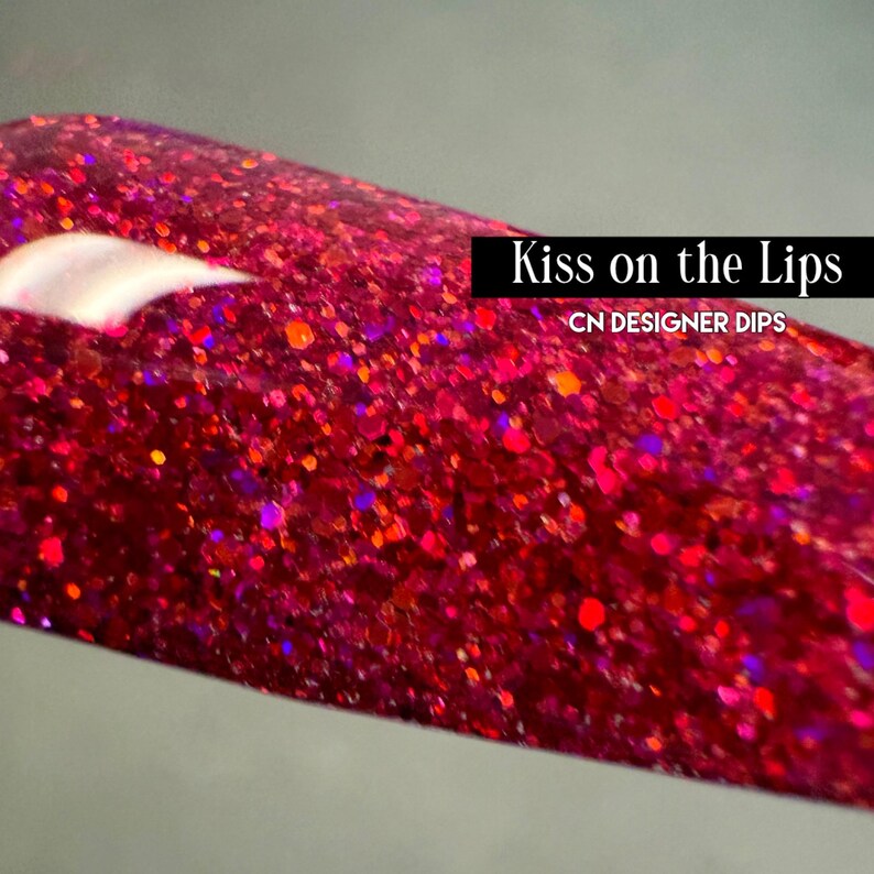 Puede incluir: Primer plano de una u&ntilde;a con un vibrante acabado rojo brillante. El esmalte de u&ntilde;as est&aacute; salpicado de purpurina roja, naranja y morada de varios tama&ntilde;os. El texto de la imagen dice "Kiss on the Lips" y "CN DESIGNER DIPS."