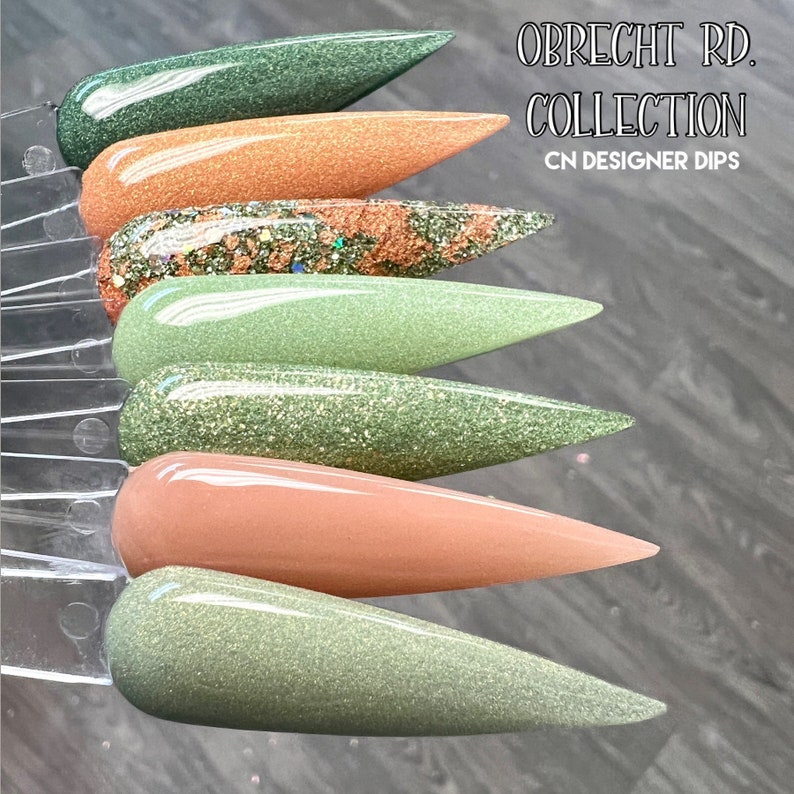 May include: A set of six nail dip powder colors in shades of green and brown. The colors are arranged in a row, with the green shades on top and the brown shades on the bottom. The set is labeled "Obrecht Rd. Collection CN Designer Dips".