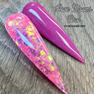 Op de afbeelding: Twee nageldippoeders, &eacute;&eacute;n roze met iriserende glitter en &eacute;&eacute;n paars. Het label luidt "Love Blooms Duo CN Designer Dips".