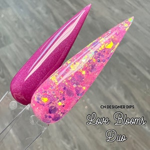 Op de afbeelding: Twee nageldipkleuren, &eacute;&eacute;n in diep roze en de andere in roze met iriserende glitter. De tekst "CN Designer Dips Love Blooms Duo" staat onder de nageldips.