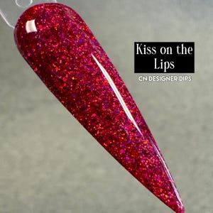 Puede incluir: Una muestra de u&ntilde;a larga y c&oacute;nica recubierta con un vibrante esmalte rojo brillante. El esmalte est&aacute; densamente lleno de part&iacute;culas brillantes, creando un efecto multitonal. El texto "Kiss on the Lips" y "CN DESIGNER DIPS" se muestra en un rect&aacute;ngulo negro.