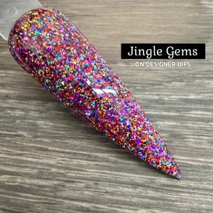 May include: A nail swatch with a glitter design. The nail is covered in a mix of red, blue, green, and gold glitter. The text "Jingle Gems" and "CN DESIGNER DIPS" are displayed on a black rectangle.