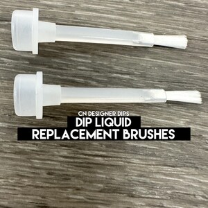 May include: Two clear plastic dip liquid replacement brushes. Each brush has a white bristle head and a long, thin stem. The text "CN DESIGNER DIPS DIP LIQUID REPLACEMENT BRUSHES" is printed in black on a black rectangle.