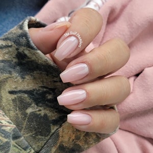 May include: Close-up of a hand with light pink square-shaped nails. The nails are polished with a sheer, light pink nail polish. The hand is resting on a camouflage fabric.