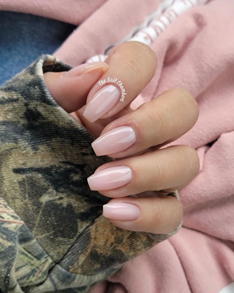 May include: Close-up of a hand with light pink square-shaped nails. The nails are polished with a sheer, light pink nail polish. The hand is resting on a camouflage fabric.