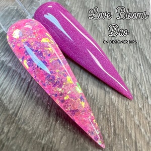 Op de afbeelding: Twee nageldippoederkleuren, &eacute;&eacute;n roze met iriserende glitter en &eacute;&eacute;n paars. De tekst "Love Blooms Duo" en "CN Designer Dips" staat op de afbeelding.