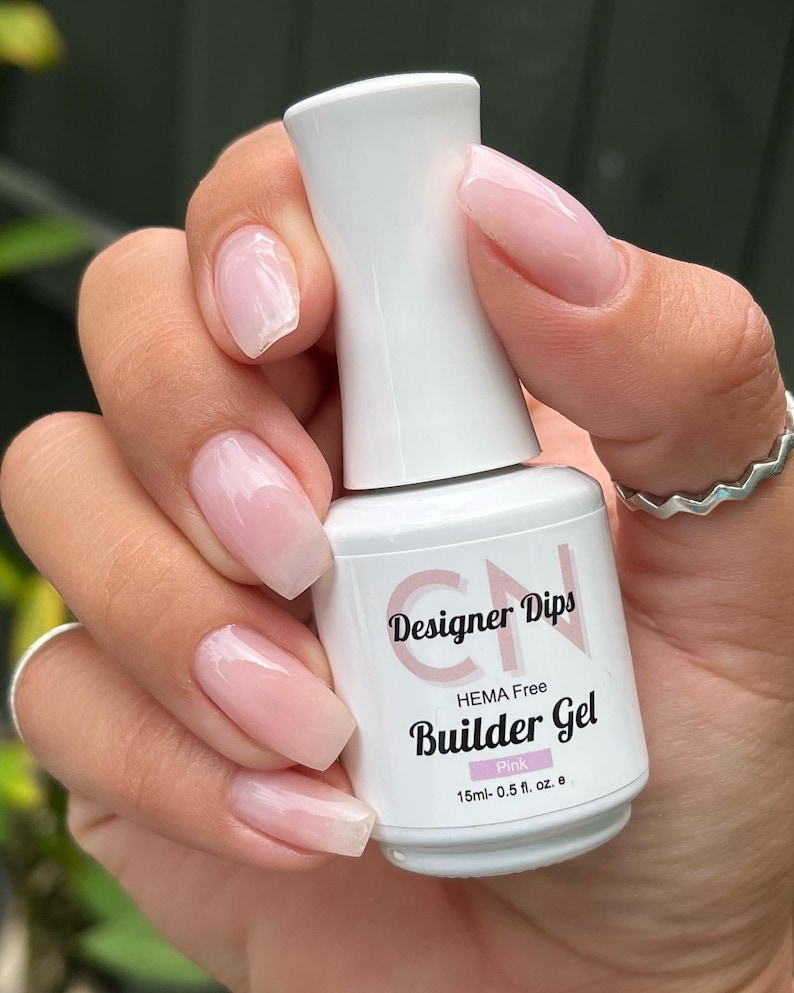 May include: A white bottle of pink builder gel nail polish with the text "Designer Dips HEMA Free Builder Gel Pink 15ml-0.5 fl. oz. e" on the label.