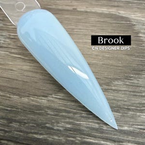 Puede incluir: Una muestra de esmalte de uñas azul claro con un acabado brillante. La muestra está etiquetada como "Brook" y "CN DESIGNER DIPS" en un rectángulo negro. La muestra se muestra sobre una superficie de madera.