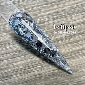May include: A close-up of a black and silver nail dip powder with a glittery finish. The nail dip powder is called "Eclipse" and is from CN Designer Dips.