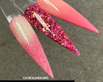 May include: Three different pink nail dip powder colors. The colors are a light pink with a shimmer, a pink with gold glitter, and a bright pink with a gradient effect. The text "CN DESIGNER DIPS" and "CRAVE COLLECTION" is at the bottom of the image.