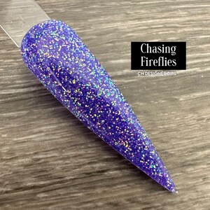 May include: A close-up of a purple nail dip powder with iridescent glitter. The powder is called "Chasing Fireflies" and is from CN Designer Dips.