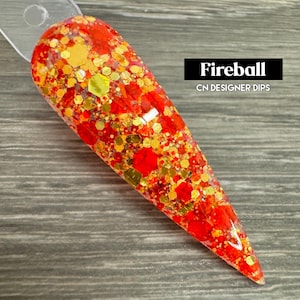 Puede incluir: Primer plano de un polvo de inmersi&oacute;n para u&ntilde;as de color rojo y dorado con purpurina. El polvo est&aacute; en un recipiente transparente y tiene una superficie texturizada. El texto "Fireball CN DESIGNER DIPS" es visible en la imagen.