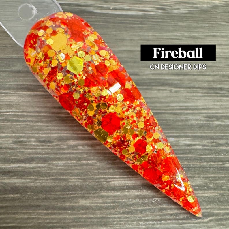Puede incluir: Primer plano de un polvo de inmersi&oacute;n para u&ntilde;as de color rojo y dorado con purpurina. El polvo est&aacute; en un recipiente transparente y tiene una superficie texturizada. El texto "Fireball CN DESIGNER DIPS" es visible en la imagen.