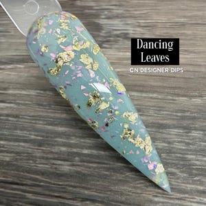 May include: A long, light blue nail with gold leaf and iridescent flakes. The nail is labeled "Dancing Leaves" and "CN DESIGNER DIPS" in a black rectangle.