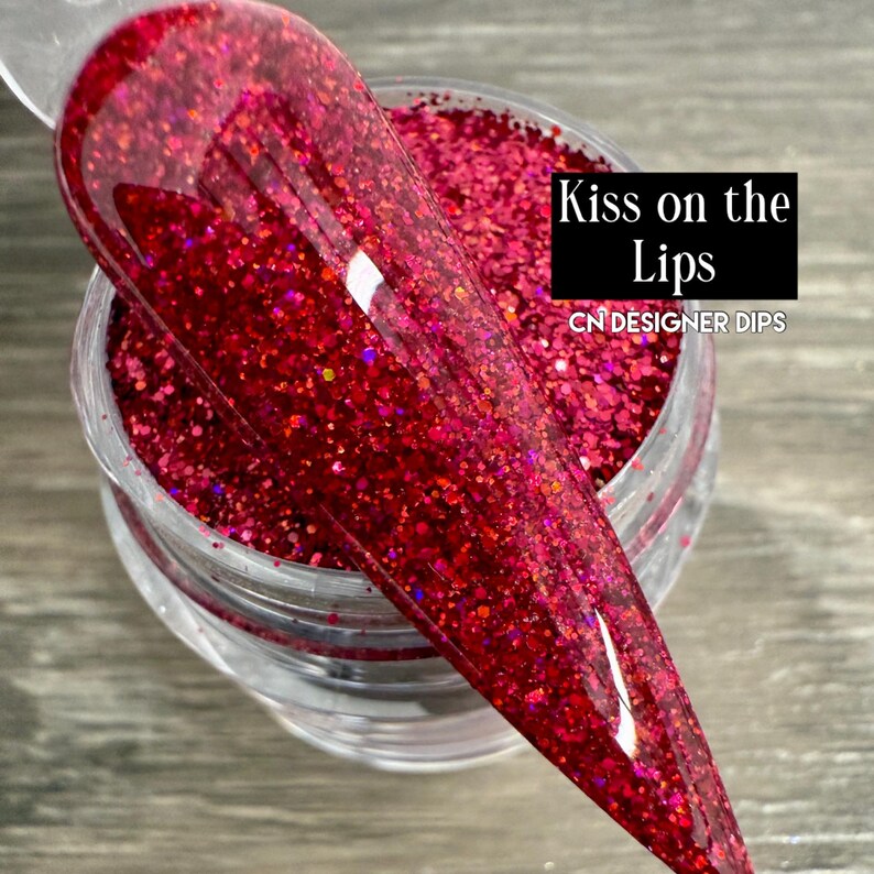 Puede incluir: Un tarro de polvo de inmersi&oacute;n para u&ntilde;as con purpurina roja con un aplicador de pl&aacute;stico transparente. El aplicador est&aacute; cubierto con la misma purpurina roja. El texto "Kiss on the Lips" y "CN DESIGNER DIPS" est&aacute; impreso en un rect&aacute;ngulo negro.