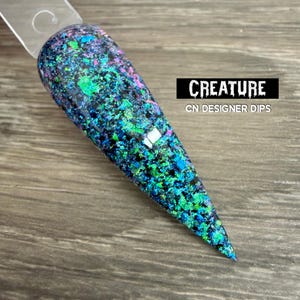 May include: A pointed nail swatch stick featuring a multi-colored glitter polish with vibrant green, blue, and purple reflective flakes over a black base. The text CREATURE and CN DESIGNER DIPS is printed in white on a black rectangular background.