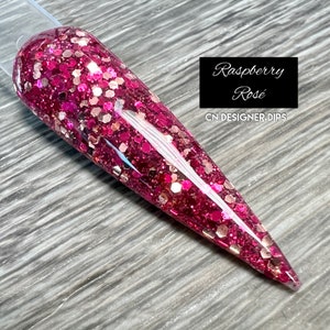 May include: A long, pointed, clear nail with a pink and gold glitter polish. The nail is labeled "Raspberry Rosé" and "CN DESIGNER DIPS".