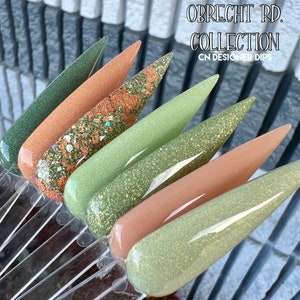 May include: A set of six nail dip powders in shades of green and brown with glitter. The set is labeled "Obrecht Rd. Collection CN Designer Dips".