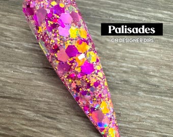 May include: A close-up of a pink, yellow, and orange glitter nail dip powder. The powder is in a long, pointed shape and is on a wooden surface. The text "Palisades CN DESIGNER DIPS" is visible in the image.