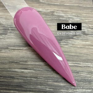 May include: A glossy, pink nail swatch with the word "Babe" in a black box. The text "CN DESIGNER DIPS" is below the word "Babe". The nail swatch is on a clear plastic stick, and the background is a wooden surface.