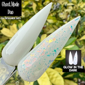 May include: Two white nail dip samples, one solid and one with iridescent glitter, are displayed against a blurred green background. The text "Ghost Mode Duo" and "CN Designer Dips" is visible. A small graphic indicates the product glows in the dark.