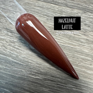 Puede incluir: Primer plano de un polvo de inmersión para uñas de color marrón con un acabado brillante. La etiqueta dice "Hazelnut Latte" y "CN Designer Dips".