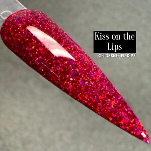Puede incluir: Una u&ntilde;a larga y puntiaguda de color rojo intenso, cubierta de purpurina, con el texto "Kiss on the Lips" y "CN DESIGNER DIPS". La u&ntilde;a tiene un acabado brillante y est&aacute; llena de part&iacute;culas brillantes. El color es un rojo rico y vibrante.
