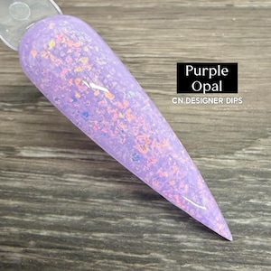 May include: A nail swatch featuring a purple opal design. The nail is a light purple hue, speckled with iridescent flakes. The text "Purple Opal" and "CN DESIGNER DIPS" is in a black rectangle.