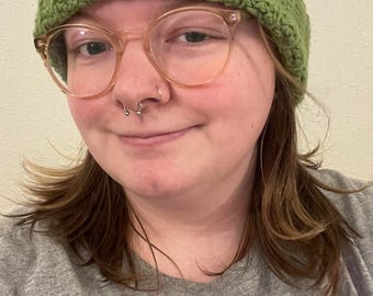 May include: A green crocheted frog hat with white and black eye details. The hat is worn by a person wearing glasses and a grey top. The hat is a fun, whimsical accessory.