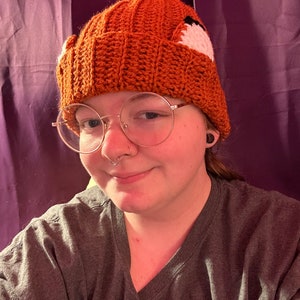 May include: An orange crochet beanie hat with fox ear details. The hat has a ribbed texture and white and black accents on the ears. A warm and cosy accessory.