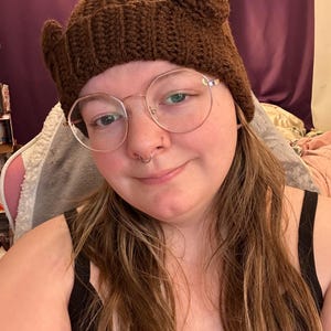 May include: A brown crocheted beanie with bear ears. The beanie is being worn by a person with long brown hair and glasses.