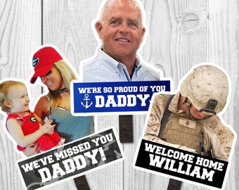 May include: Three photo booth props with a military theme. The first prop features a man's face with the text "We're so proud of you Daddy". The second prop features a woman holding a child with the text "We've missed you Daddy!". The third prop features a soldier with the text "Welcome home William".