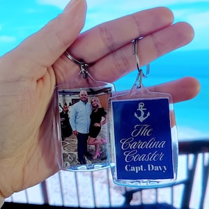 May include: Two clear plastic keychains with silver rings. One keychain has a photo of a couple in front of a building. The other keychain has a blue background with a silver anchor and the text "The Carolina Coaster Capt. Davy".