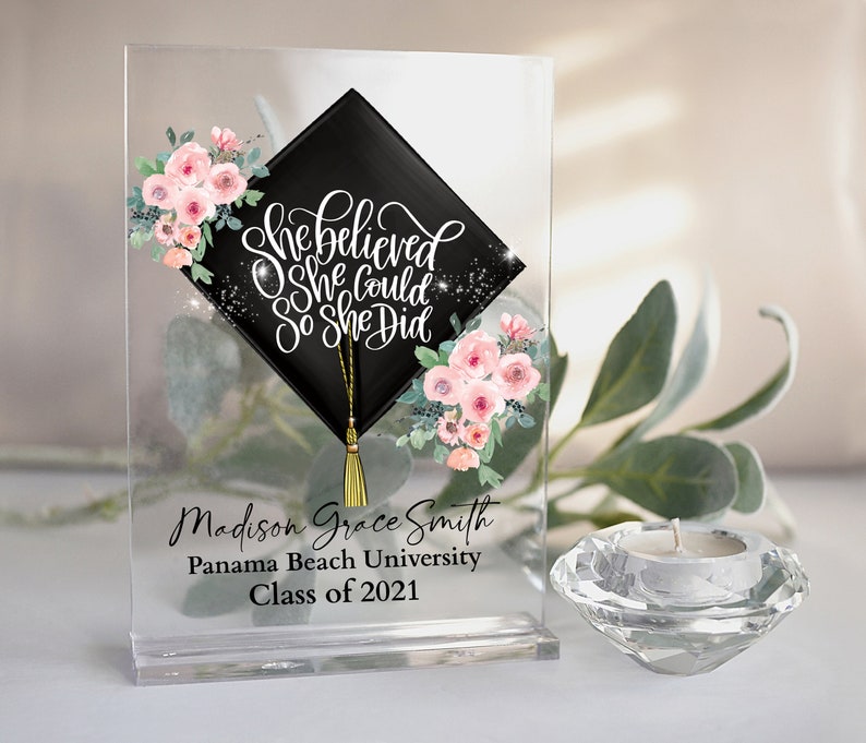 Puede incluir: Recuerdo de graduaci&oacute;n de acr&iacute;lico transparente con un dise&ntilde;o de birrete negro y el texto "She Believed She Could So She Did". Presenta detalles florales y personalizados. Incluye un portavelas de cristal.
