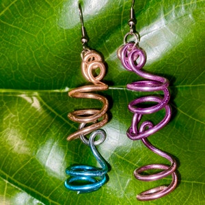 Puede incluir: Un par de pendientes hechos a mano con un diseño en espiral. Los pendientes están hechos de alambre y tienen un degradado de colores, comenzando con un rosa claro en la parte superior y desvaneciéndose a un azul más oscuro en la parte inferior.