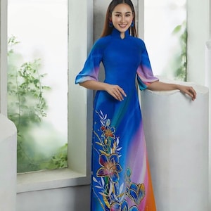 May include: A woman wearing a blue and orange long-sleeved dress with a floral print. The dress has a mandarin collar and a fitted silhouette. The woman is standing in front of a window with a white frame.