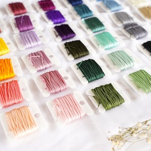 Könnte beinhalten: Eine Auswahl an bunten Stickgarnrollen, die in Reihen auf einer weißen Oberfläche angeordnet sind. Das Garn ist in einem Regenbogenmuster angeordnet, mit Schattierungen von Rot, Orange, Gelb, Grün, Blau, Lila und Braun.