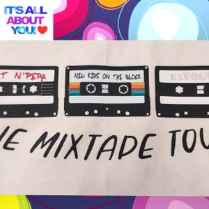 May include: A white tote bag featuring three black cassette tapes printed on it. The tapes have the names "T N'PERA", "NEW KIDS ON THE BLOCK", and "ENVOG" printed on them. The text "THE MIXTAPE TOUR" is printed below the tapes.