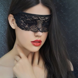 Könnte beinhalten: Eine Frau mit langen schwarzen Haaren trägt eine schwarze Spitzen-Augenmaske. Sie trägt ein schwarzes Top mit offener Schulter und ihre Hände sind vor ihr gefaltet.