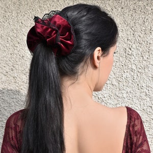 May include: A deep red velvet hair scrunchie featuring a delicate black lace trim along the outer edge. The fabric has a soft, reflective sheen, and the accessory is gathered into a voluminous, circular shape to secure a ponytail of dark hair.