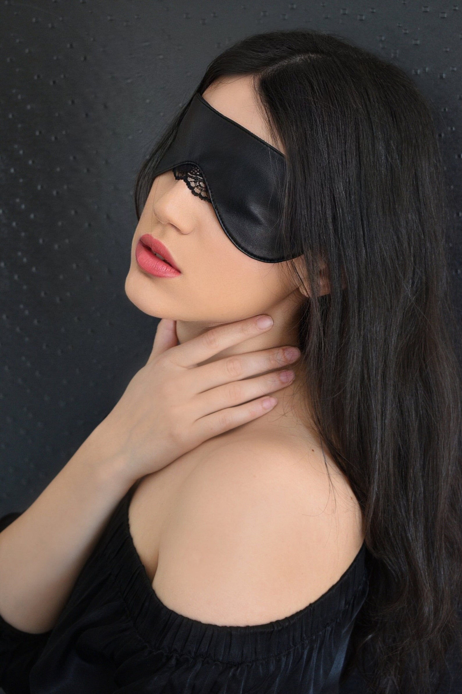 Woman wearing black leather blindfold mask with lace trim, hand on neck, evoking sensual and mysterious mood.