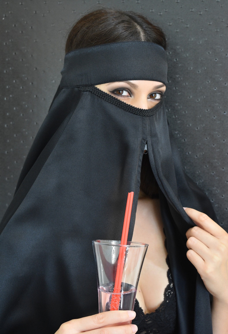 Peut inclure: Une femme portant un niqab noir avec une paille rouge dans un verre de liquide rose.