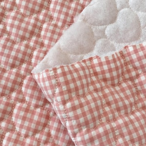 May include: Close-up of a quilted fabric with a heart pattern. The fabric features a gingham pattern in white and light red. The hearts are outlined with white stitching, and the backing is a soft white material.