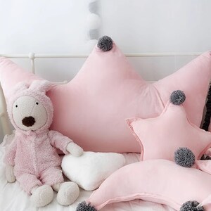 May include: Pink star-shaped pillow with gray pom-poms, a pink crescent moon pillow, and a white heart-shaped pillow. The pillows are on a white bed with a pink stuffed animal.