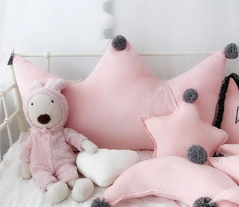May include: Pink star-shaped pillow with gray pom-poms, a pink crescent moon pillow, and a white heart-shaped pillow. The pillows are on a white bed with a pink stuffed animal.