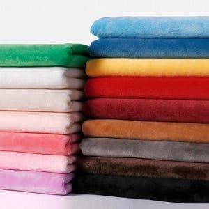 Può includere: Pile di tessuto in peluche in vari colori, tra cui verde, bianco, rosa e viola a sinistra, e blu, giallo e rosso a destra. Il materiale morbido è piegato con cura, mostrando una gamma di tonalità.