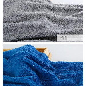 May include: Two fabric swatches, one gray and one blue, are shown on a wooden surface. The swatches are soft and fluffy, with a fuzzy texture. The number "11" is written on a white label below the gray swatch, and the number "12" is written on a white label below the blue swatch.